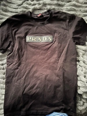 Svart t-shirt från Prada - Säljer en svart t-shirt från Prada med stort logotyp-tryck på bröstet. Klassisk rund halsringning och korta ärmar. Tillverkad i mjuk bomull för en skön känsla och enkel stil.