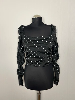 Superfin svart prickig blus H&M oanvänd - Supersnygg svart blus från H&M med vita prickar, puffiga långärmar och rynkade detaljer både fram och bak. Blusen har en fyrkantig ringning och dragkedja i ryggen. Materialet är glansigt och känns lyxigt, perfekt för att sticka ut lite extra.