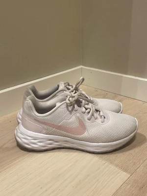 Vita Nike sneakers med rosa swoosh - Säljer ett par fräscha vita Nike sneakers med ljusrosa swoosh på sidan. Skorna har snörning, mesh-ovandel för bra andningsförmåga och en chunky, mönstrad sula som ger grym komfort. Perfekta till träning eller när du vill ha en sportig look.