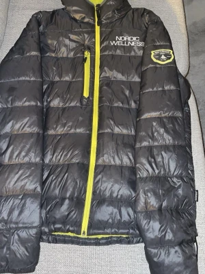 Svart pufferjacka från Matterhorn  - Säljer en svart pufferjacka från Matterhorn med gul insida och detaljer. Jackan har huva, dragkedja i gult, bröstficka och broderad patch på ärmen. På bröstet står det 'Nordic Wellness'. Perfekt för kalla dagar och skidåkning. 