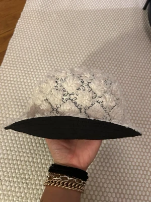 Reversibel bucket hat från Lala Berlin - Säljer en unik bucket hat från Lala Berlin som är vändbar. Ena sidan är svart i slätt material med broderad logga, andra sidan är fluffig vit med rutigt mönster och svart text. Perfekt accessoar för att lyfta din look.