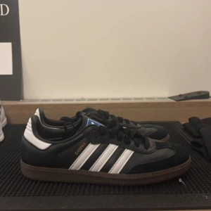 Adidas Samba svart/vit sneakers - Klassiska Adidas Samba sneakers i svart skinn med vita detaljer och de ikoniska tre ränderna på sidorna. Skorna har brun gummisula, svart snörning och blå Adidas-logga på plösen. Perfekt för dig som gillar retrostil och streetwear.( Äkta morsan jobbar där)