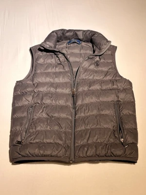 Grå dunväst från Polo Ralph Lauren - Säljer en grå dunväst från Polo Ralph Lauren i storlek S. Västen har hög krage, två sidofickor med dragkedja och quiltad design. Perfekt att slänga över en hoodie eller tröja för extra värme. Klassisk och stilren look. Hundra procent äkta 💯 