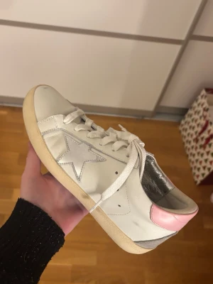 Golden goose skor - Snygga vita sneakers i skinn med en metallic-silver stjärna på sidan och en ljusrosa hälkappa. Skorna har vita snören, rund tå och platt sula. Perfekta för dig som vill ha en clean men ändå unik look med lite extra detaljer.