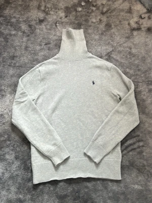Ljusgrå polotröja från Ralph Lauren - Säljer en stilren ljusgrå polotröja från Ralph Lauren med klassisk broderad logga på bröstet. Tröjan är stickad i mjuk bomull, har lång ärm och ribbade muddar samt hög polokrage. ny pris 2000kr/ mitt pris 600kr