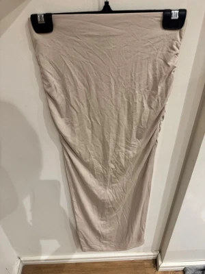 Beige långkjol i mjukt material XS - Säljer en beige långkjol i mjukt och stretchigt material. Kjolen har en enkel, rak passform och lätt rynkade detaljer längs sidorna för en snygg siluett. Perfekt att styla med en crop top eller oversized tröja.
