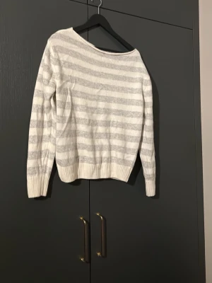 Randig vit och grå stickad tröja - Mysig stickad tröja i vitt och ljusgrått med horisontella ränder. Tröjan är off shoulder och har långa ärmar samt ribbade muddar vid ärmslut och nederkant. Nypris 600kr och endast använd två gånger!