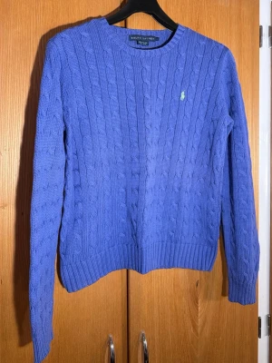 Ralph Lauren Blå kabelstickad tröja   - Kabelstickad blå tröja från Ralph Lauren i 100% bomull. Klassisk modell med rund halsringning, ribbade muddar och det ikoniska broderade logotypen på bröstet. Perfekt för lager-på-lager och chill stil.