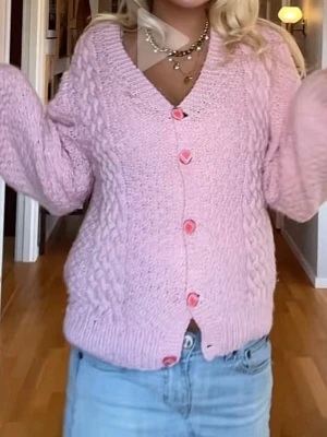 Ljusrosa stickad kofta med knappar - Supersöt ljusrosa stickad kofta med flätstickade detaljer och v-ringning. Koftan har långa ärmar och rosa knappar framtill som ger en gullig touch. Perfekt att slänga över en topp för en mysig look.
