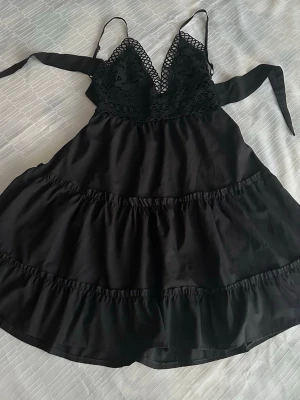 Svart klänning med knytband M - Säljer en svart väst med tunna axelband och breda knytband i sidan. Överdelen har detaljerad spets med blommönster och v-ringad form. Nederdelen är i slät polyester med volang. Perfekt för att lyfta en outfit med en edgy touch.