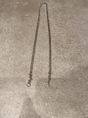 Halskedja i silverfärg - Säljer en stilren kedja i silverfärgad.Kedjan har kraftiga länkar och två karbinhakar i ändarna för enkel fastsättning. Längden är cirka 60 cm. Perfekt accessoar för att ge din outfit en cool touch.