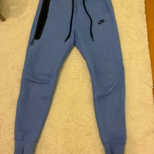 Nike Tech mjukisbyxor, nya - Snygga ljusblå mjukisbyxor från Nike med svarta snören i midjan och en svart detalj vid fickan. Byxorna har ribbade muddar vid bensluten och Nike-logga på låret.  Storlek S och inköpta i footlocker. Endast testade