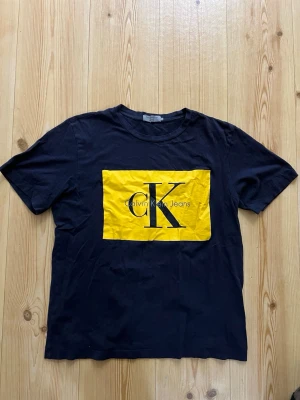 Svart t-shirt Calvin Klein Jeans L - Snygg svart t-shirt från Calvin Klein Jeans i storlek L. Framsidan har en stor gul ruta med CK-logga och text. Klassisk passform och rund hals. Tillverkad i mjuk bomull som känns skön mot huden. Perfekt för dig som gillar stilrena och trendiga plagg.