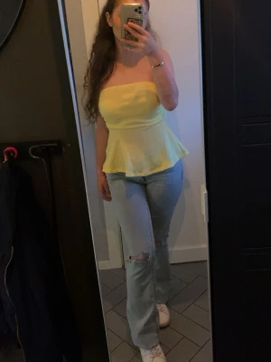 Gul bandeau tubtopp med volang - Säljer en söt gul bandeau tubtopp med volang nertill. Toppen är axelbandslös och har en lätt utsvängd form som ger en feminin siluett. Perfekt för varma dagar och enkel att styla med jeans eller kjol. Den är lite genomskinlig, det beror på vad man har under. 