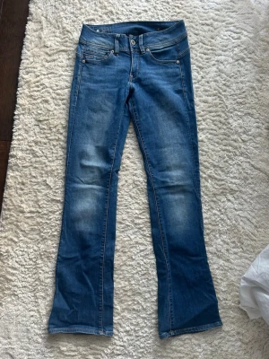 Bootcut jeans  - Blå bootcut-jeans i denim från G-Star RAW, storlek W27 L32. Smickrande bootcut-passform med lätt stretch för extra komfort.  ✔ Mycket sparsamt använda ✔ Inga defekter eller skador ✔ Unik design baktill ✔ Diskret brodyr på bakfickorna ✔ Högkvalitativ denim