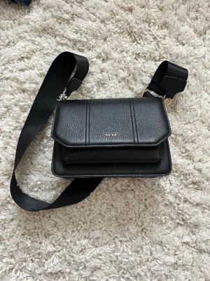 Svart axelväska från NA-KD med ormmönster - En svart axelväska från NA-KD i skinnimitation med bred axelrem och guldfärgade detaljer, sidorna har ett ormmönsher. Säljer så den tyvärr inte kommer till användning, använd fåtal gånger!