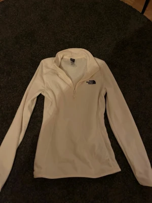 Vit fleece-tröja från The North Face - Säljer en vit långärmad fleece-tröja från The North Face med halv dragkedja framtill. Tröjan har en clean look med logga på bröstet och är perfekt för kyliga dagar. Slimmad passform och mjukt material som är skönt mot huden.