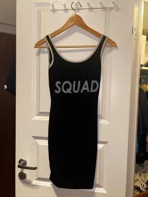Svart kortklänning från Boohoo - Säljer en svart tight kortklänning från Boohoo med vita kanter och texten 'SQUAD' framtill. Klänningen är ärmlös och har en sportig look med rundad halsringning. Perfekt för dig som vill sticka ut med en enkel men cool stil.