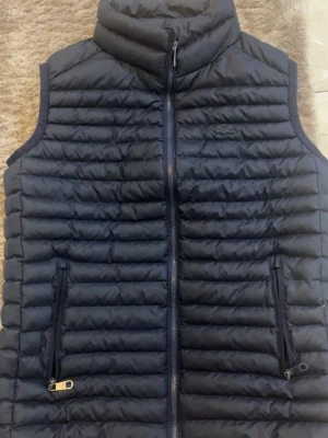 Mörkblå dunväst från Lacoste - Säljer en mörkblå dunväst från Lacoste med quiltad design och dragkedja framtill. Västen har två fickor med dragkedja och diskret Lacoste-logga på bröstet. Insidan har en innerficka med dragkedja och elastiska kanter med Lacoste-tryck.