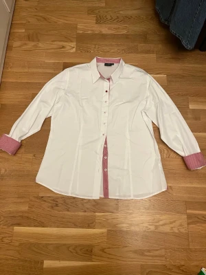 Vit skjorta Ernst Alexis rutig detalj - Klassisk vit skjorta från Ernst Alexis med rödrutiga detaljer på krage, knappslå och manschetter. Långärmad modell i bomull med normal passform och knappar framtill. Perfekt för dig som gillar stilrena plagg med en twist.