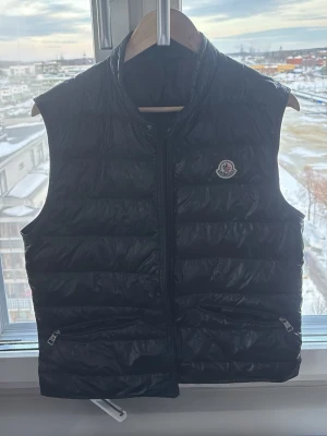Svart dunväst från Moncler - Säljer en svart dunväst från Moncler med klassisk quiltad design och hög krage. Västen har dragkedja framtill, två fickor med dragkedja och Moncler-logga på bröstet. Perfekt för lager-på-lager och snygg till både hoodie och t-shirt. En perfekt väst till dig som vill sticka ut lite! Dragkedjan är lite konstig men det går att knäppa den om man försöker och vill! Skriv om ni har frågor. Kan självklart går ner i pris om behövelse