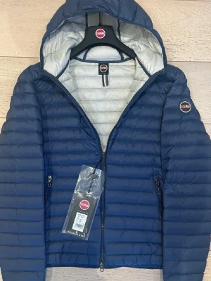 Blå dunjacka från Colmar med huva - Snygg blå dunjacka från Colmar med quiltad design och huva. Jackan har dragkedja framtill, två sidofickor med dragkedja och en ljus insida. Perfekt för kyliga dagar och enkel att matcha med olika outfits. Plats till ettik är borta