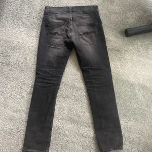 Nudie Jeans Grim Tim Concrete Black - Säljer ett par svarta Nudie Jeans Grim Tim i färgen Concrete Black. De har en slim passform med raka ben och klassisk femficksdesign. Tillverkade i 99% ekologisk bomull och 1% elastan för extra stretch. Snyggt slitna detaljer och diskret logga på bakfickan.har en defekt längst ner men inget märkvärdigt vid användning 
