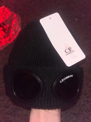 Svart mössa med goggles från C.P. Company - Säljer en svart ribbstickad mössa från C.P. Company med ikoniska goggles framtill. Mössan är tillverkad i 100% ull och har en snygg, modern look med tydlig logga på glasögonen. Perfekt accessoar för dig som vill sticka ut. Fraktar samma dag👍😁