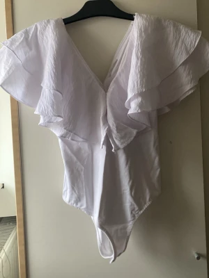 Vit volangbody från Zara - Säljer en vit volangbody från Zara med djup v-ringning och  volangdetaljer över axlarna. Bodyn är i ett lätt material och har en feminin, flowy look som passar perfekt till olika outfits. Supersnygg att styla med både jeans och kjol. Endast använd 1 gång 