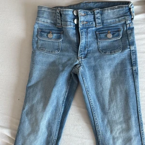 Ljusblå bootcut jeans med fickor - Säljer ett par ljusblå bootcut jeans med dubbla knappar i midjan och två snygga fickor framtill med lock och knapp. Jeansen har breda benslut och klassisk jeanslook. Materialet är en mix av bomull och polyester, vilket gör dem både mjuka och slitstarka.