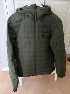 Skidset från J.Lindeberg M - Skidkläder från J.Lindeberg i storlek M. Jackan är olivgrön och har huva, dragkedja framtill och diskret logga på bröstet samt ärmen. Snygg quiltad design och praktiska fickor. Svarta skidbyxor i strl M!