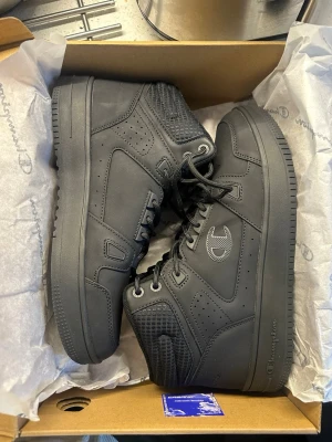Svarta höga sneakers från Champion - Säljer ett par svarta höga sneakers från Champion med snyggt broderad logga på sidan. Skorna har snörning, vadderad och ribbad plös samt detaljer i syntetmaterial. Perfekta för dig som gillar streetwear och vill ha en clean look.