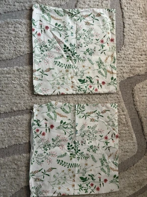 Blommiga kuddfodral i bomull H&M - Säljer ett par söta kuddfodral i vit bomull med blommigt mönster i grönt, rosa och beige. Kuddfodralen stängs med dragkedja. På ena kuddfodralet har sömmen öppnat sig cirka 10cm. (På sista bilden) 50x50 