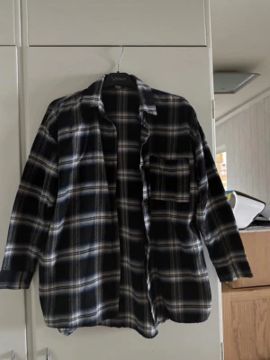 Oversized rutig skjorta från YesStyle - Säljer en oversized rutig skjorta från YesStyle i svart, blått och vitt. Skjortan har klassisk krage, bröstficka och knappar framtill. Perfekt för dig som gillar en avslappnad och trendig stil.