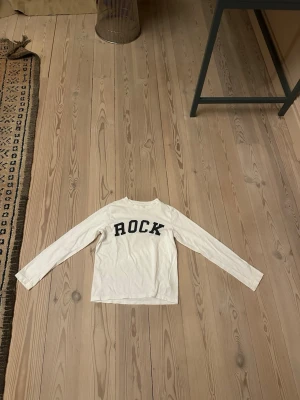 Vit långärmad topp Zadig & Voltaire - Säljer en vit långärmad topp från Zadig & Voltaire med svart text 'ROCK' framtill. Klassisk rak passform och rund halsringning. Tillverkad i mjuk bomull som känns skön mot huden. Perfekt för dig som gillar en enkel men cool stil.10år