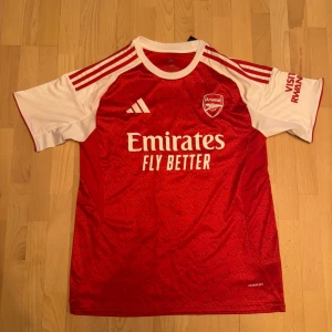 Arsenal matchtröja Gyökeres 14 Adidas - Säljer en officiell Arsenal matchtröja från Adidas med Gyökeres och nummer 14 på ryggen. Tröjan är röd ymed vita ärmar, har vita Adidas-ränder på axlarna och klubbmärke på bröstet. Materialet är lätt polyester med Aeroready-teknologi och snyggt mönster i tyget.