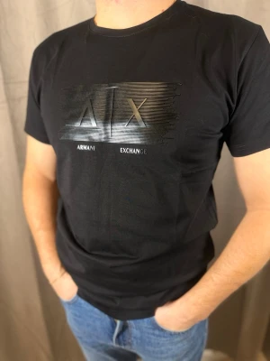 Svart Armani Exchange t-shirt - Snygg svart t-shirt från Armani Exchange med ett stilrent, glansigt AX-tryck framtill. Klassisk rund halsringning och korta ärmar. Tillverkad i mjuk bomull som känns skön mot huden. Perfekt för dig som gillar en clean och modern look.