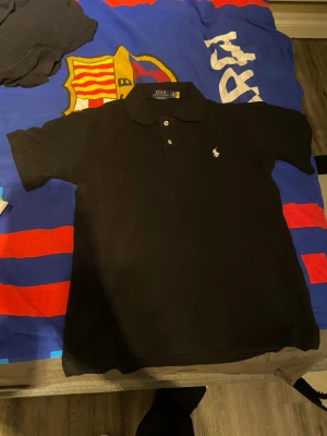 Svart pikétröja från Ralph Lauren - Klassisk svart pikétröja från Polo Ralph Lauren i modellen Custom Slim Fit. Tröjan har korta ärmar, krage med två knappar och den ikoniska broderade loggan i vitt på bröstet. Tillverkad i mjuk bomull som känns skön mot huden.