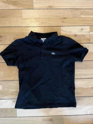 Svart pikétröja från Lacoste - Klassisk svart pikétröja från Lacoste med korta ärmar och krage. Tröjan har två knappar framtill och den ikoniska gröna krokodilloggan broderad på bröstet. Tillverkad i bomull för en skön och luftig känsla.