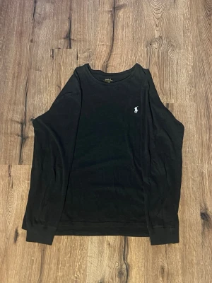 Svart långärmad tröja Polo Ralph Lauren - Klassisk svart långärmad tröja från Polo Ralph Lauren med den ikoniska vita polospelaren broderad på bröstet. Tröjan har rund halsringning och är gjord i mjuk bomull, perfekt för en clean och stilren look.