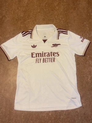 Arsenal x Adidas Originals – Tredjetröja (24/25)  - Säljer nu denna stilrena Arsenal tredjetröja för säsongen 2024/25. Tröjan kombinerar en klassisk off-white bas med eleganta detaljer i vinrött och guld.