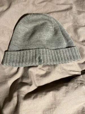 Grå stickad mössa från Polo Ralph Lauren - Snygg grå stickad mössa från Polo Ralph Lauren med klassisk ribbad kant och broderad logga framtill. Perfekt accessoar för kyliga dagar och enkel att matcha med olika outfits. Har använts för ett par år sedan men bara legat hemma helt oanvänd. Storleken är one size, 46cm. Pris går att diskutera