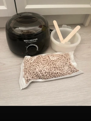 Vaxvärmare med tillbehör - Säljer en svart elektrisk vaxvärmare med genomskinligt lock, metallinsats, 2 st silikonbehållare och 2 träspatlar. Medföljer även ett paket vaxpärlor med doft av kokos. Perfekt för enkel hårborttagning hemma. Allt du behöver för smidig vaxning ingår.