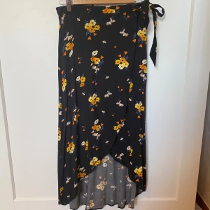 Svart blommig omlottkjol med knytband - Svart långkjol med blommigt mönster i gult, vitt och orange. Kjolen har omlottmodell med knytband i midjan och faller snyggt med en lätt volangkant framtill. 💕