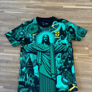 Nike Brasil grafisk fotbollströja M | Kristusmotiv & färgstarka detaljer - .Säljer en unik Brasilien-fotbollströja från Nike, storlek M, med ett grafiskt tryck av Kristusstatyn och brasilianska motiv i grönt, svart och gult. Tröjan har kortärmad modell, rund hals och är tillverkad i lätt, ventilerande polyester – perfekt för match, träning eller streetwear.  ✔️ Lätt och andas ✔️ Kortärmad, regular fit ✔️ Aldrig använd  Storlek: M Skick: Ny / aldrig använd 
