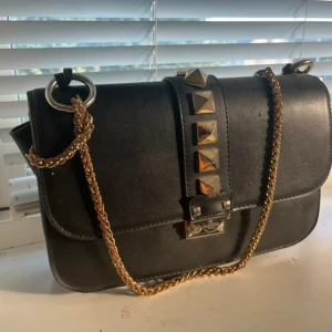 Svart axelväska ”Valentino” med nitar - Snygg svart axelväska från Valentino i skinn med guldfärgad kedja och stora pyramidformade nitar på framsidan. Väskan har ett innerfack med dragkedja och röd insida. Perfekt för dig som vill ha en edgy och lyxig accessoar.  