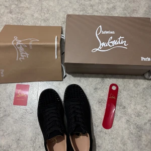 Svarta Christian Louboutin sneakers - Säljer ett par svarta sneakers från Christian Louboutin med ikoniska röda sulor och svarta nitar på tån. Skorna har snörning och är tillverkade i skinn med en platt sula. Kommer med originalkartong, påse och skohorn.