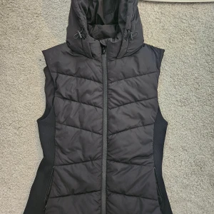 Svart dunväst med huva från Zara - Svart quiltad dunväst från Zara med dragkedja framtill och stor huva med dragsko. Västen har en figurnära passform och ribbade sidor för extra komfort. Perfekt för lager-på-lager under kyliga dagar.