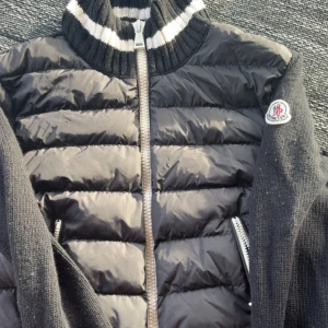 Svart pufferjacka från Moncler - Säljer en svart pufferjacka från Moncler med stickade ärmar och krage. Jackan har dragkedja framtill, två fickor med dragkedja och en randig detalj i kragen. Klassisk Moncler-logga på armen. 