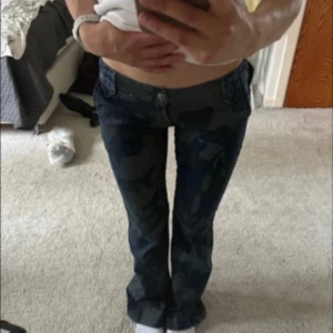 Camo bootcut jeans med fickor - Snygga camouflage-mönstrade jeans med bootcut passform och låg midja. Byxorna har stora fickor bak med knappdetalj och är i ett robust jeanstyg. Perfekta för dig som vill sticka ut med en cool streetstil. Köpta för 700, de är i nyskick med lappen kvar!